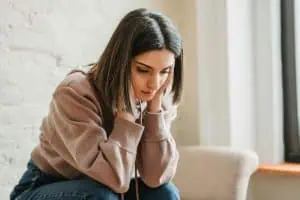 Young woman sitting indoors with a worried expression, holding her face in both hands—depicting emotional stress or anxiety.
