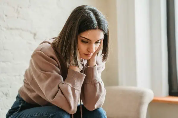 Young woman sitting indoors with a worried expression, holding her face in both hands—depicting emotional stress or anxiety.