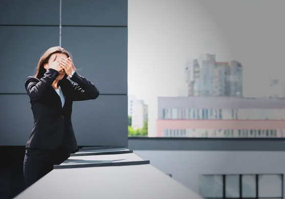 A woman in a black business suit stands on a rooftop covering her face with both hands, expressing overwhelming stress or emotional distress against a backdrop of city buildings.