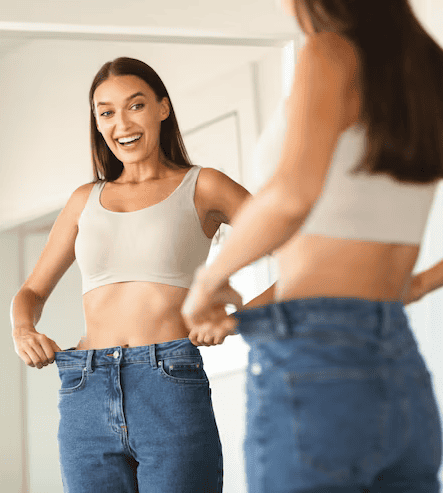 Successful slimming concept excited caucasian lady wearing jeans after weight loss comparing size posing near mirror