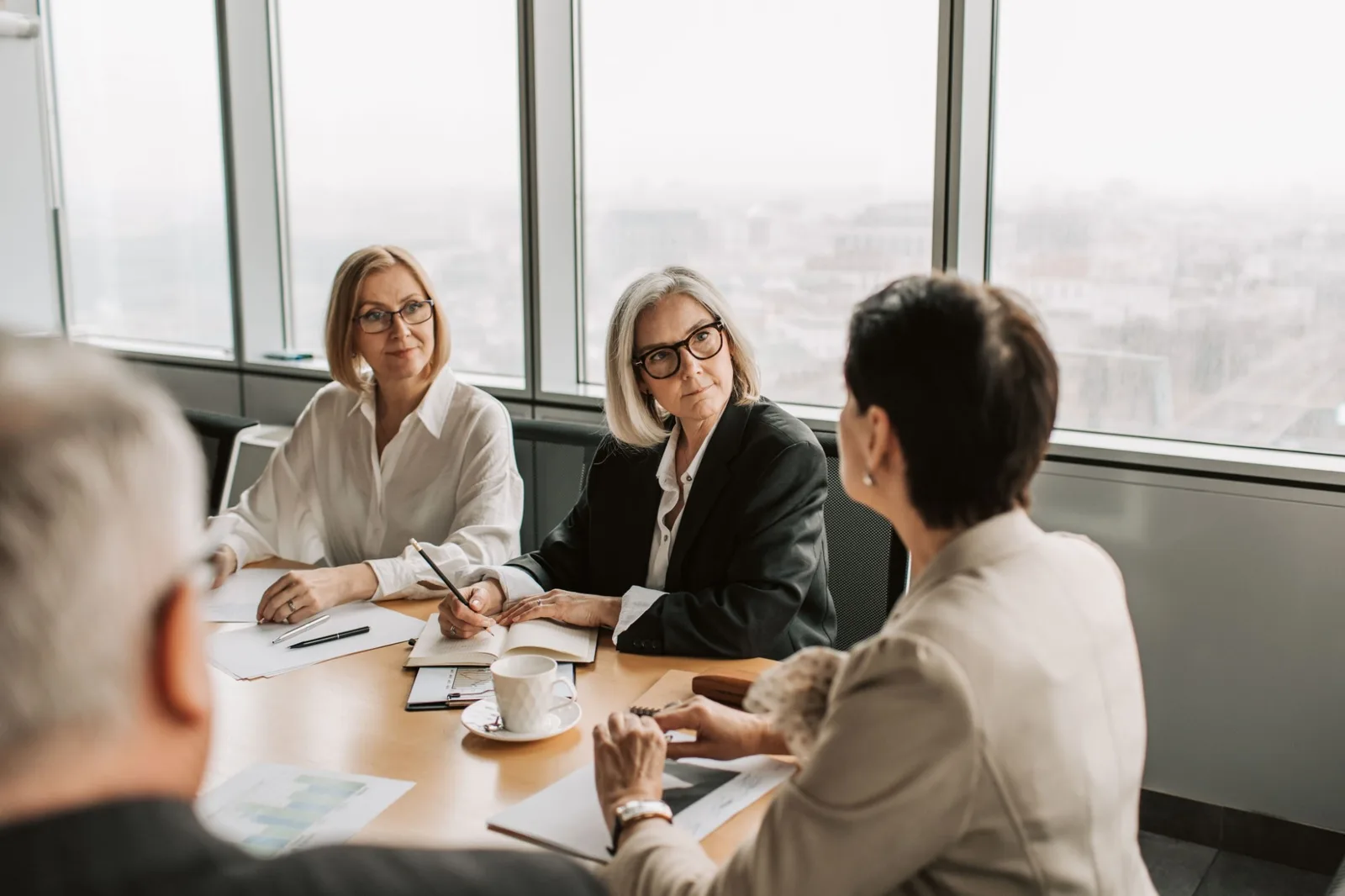 Four colleagues in a meeting around a boardroom table, two of them listening as a colleague speaks, with a city view through the windows behind them
