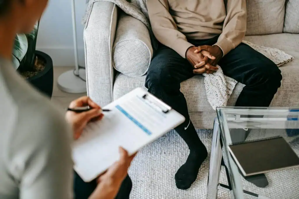 Teen sitting on a couch during a therapy session, with a counselor taking notes on a clipboard across from them.