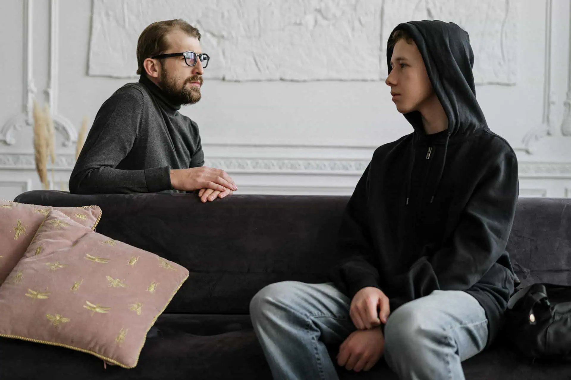 Male therapist calmly talking with a teenage boy in a hoodie during a one-on-one counseling session on a couch.