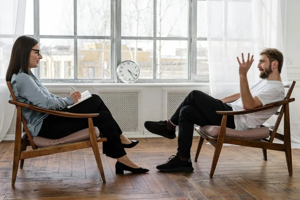 Male client speaking with a female therapist during a counseling session in a bright, modern office setting.