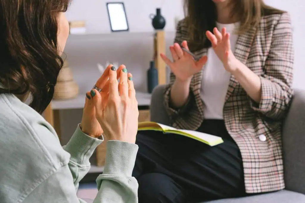 Two women in a therapy or counseling session, seated and engaged in conversation with expressive hand gestures, one holding an open notebook.