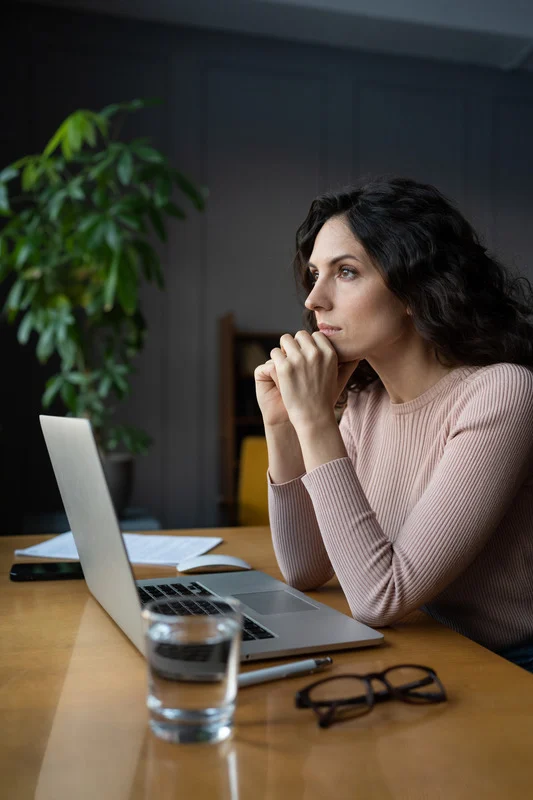 A woman sits at a desk with her hands clasped in front of her mouth, looking thoughtfully at a laptop screen. She appears deep in concentration or contemplation, with a glass of water, notebook, and glasses on the table.
