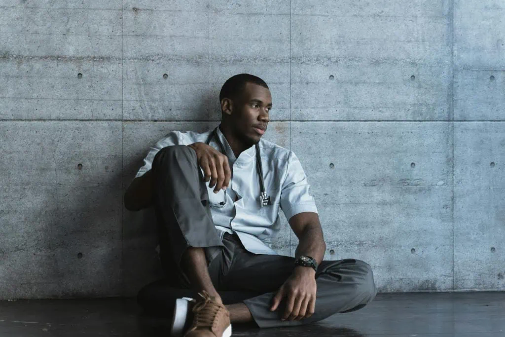 A tired healthcare worker in scrubs and a mask sits on the floor against a wall with eyes closed and head tilted back, portraying physical and emotional exhaustion from long hours and demanding shifts.