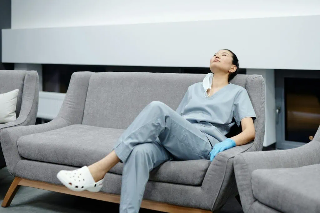 A tired shift worker in light blue scrubs rests on a gray couch with their head tilted back. A face mask hangs loosely around their neck, and they wear a blue glove on one hand and white clogs on their feet. The setting appears to be a hospital or clinic break room.