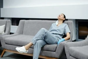 A tired shift worker in light blue scrubs rests on a gray couch with their head tilted back. A face mask hangs loosely around their neck, and they wear a blue glove on one hand and white clogs on their feet. The setting appears to be a hospital or clinic break room.