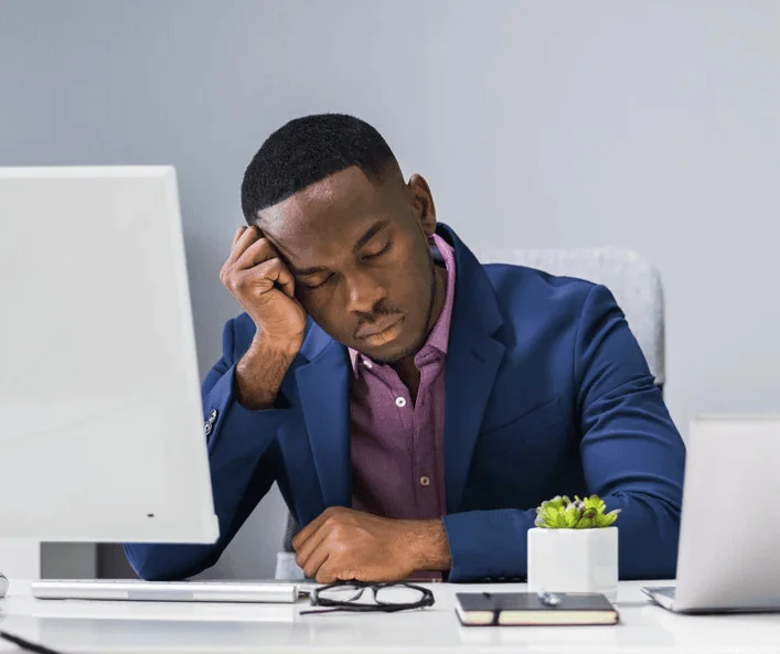 A man in a business suit sits at a desk with his head resting on his hand, eyes closed, appearing exhausted or asleep in front of a computer, suggesting fatigue or workplace burnout.
