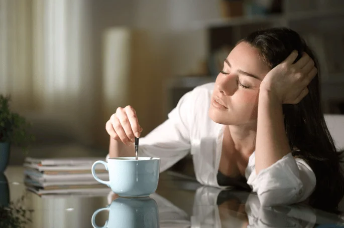 A sleepy woman in a white blouse stirring her coffee with a tired expression, resting her head on her hand, possibly struggling to wake up in the morning.