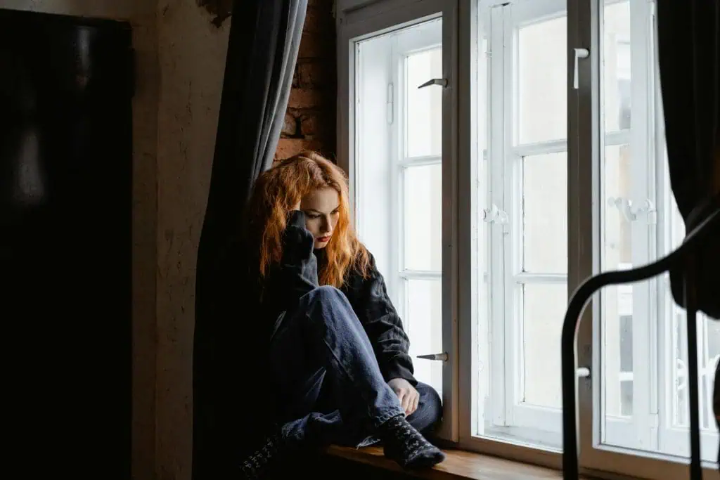 A woman sitting by the window appearing emotionally distressed, possibly experiencing TMJ disorder in women.
