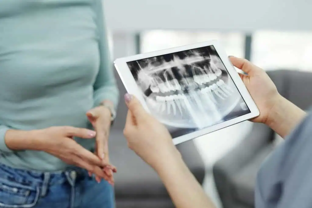 A dentist holding a tablet displaying a dental X-ray while discussing results with a patient, possibly reviewing signs of TMJ disorder.