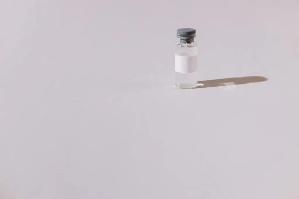 A small glass vial with a blank white label and gray cap, standing on a pale surface with a soft shadow.