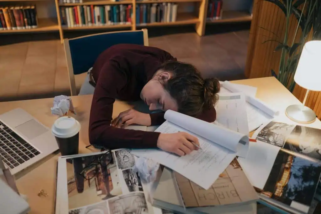 Exhausted woman asleep on a cluttered desk with papers, books, and a laptop, showing signs of burnout and mental fatigue.