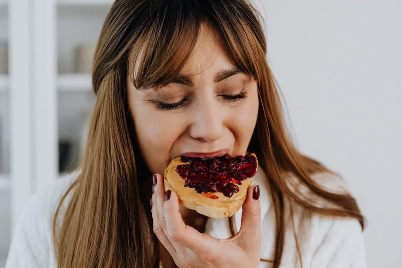 A woman takes a bite of a slice of bread topped with peanut butter and berry jam, enjoying a simple and classic snack with her eyes closed in satisfaction.