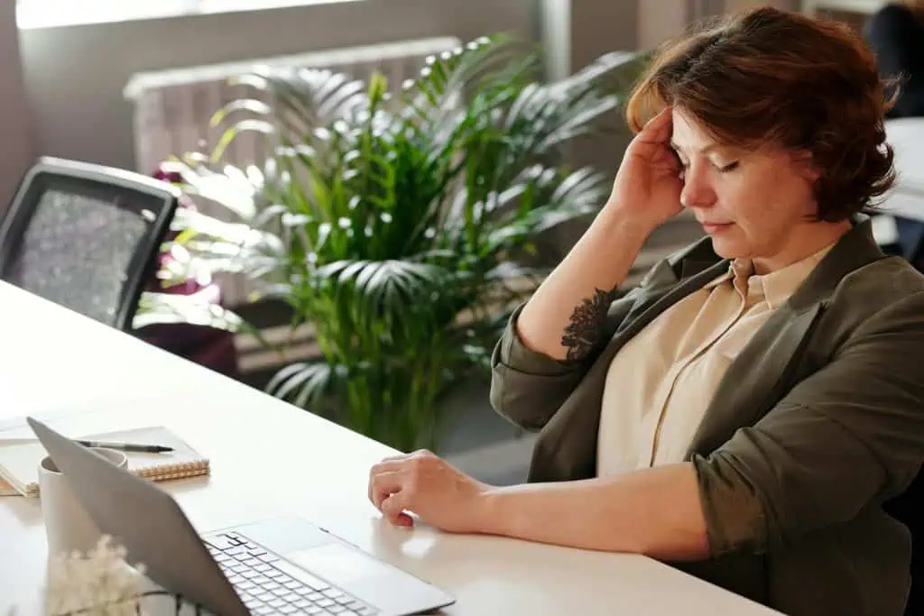Woman sitting at a desk with eyes closed, holding her head in discomfort, showing signs of stress or a tension headache.