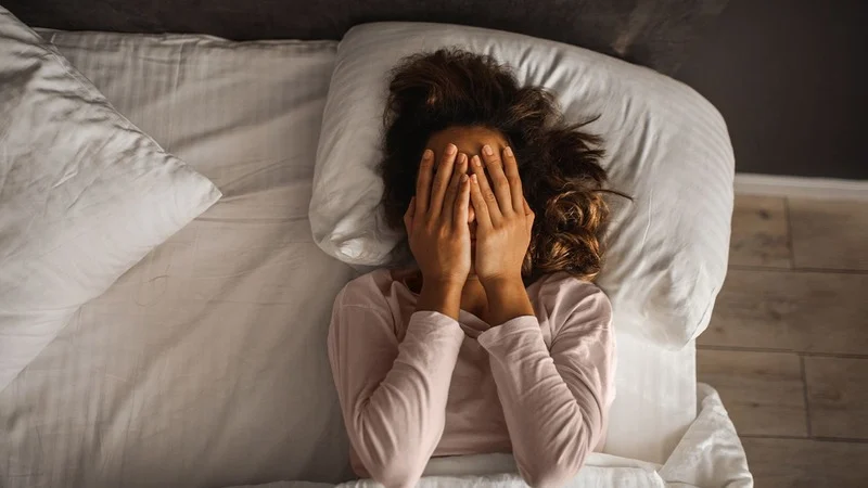 A woman lies in bed with her hands covering her face, conveying feelings of stress, anxiety, or emotional exhaustion, as she rests on white pillows in a dimly lit bedroom.