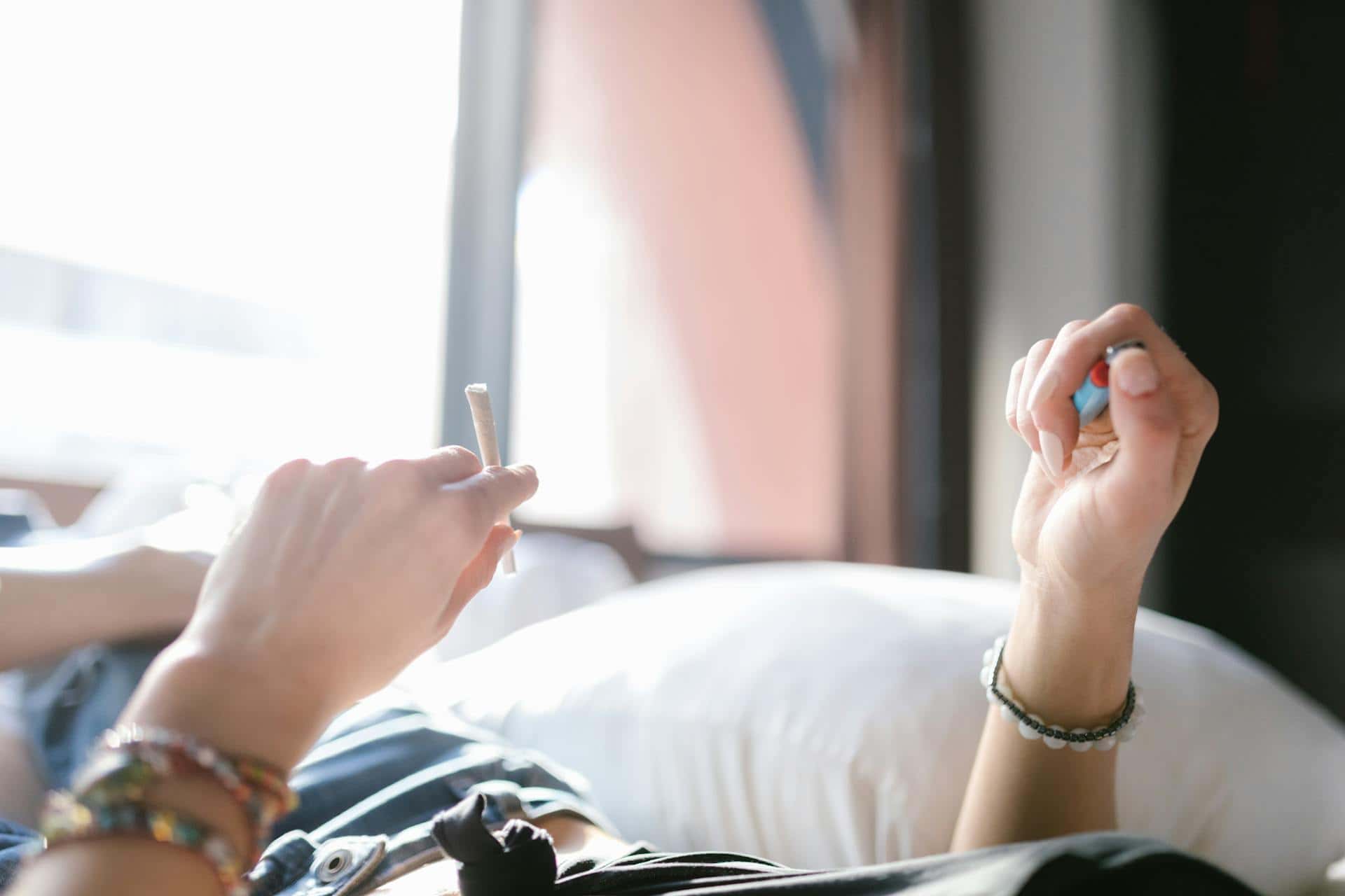 Two hands raised above a bed, one holding a rolled cigarette and the other a lighter, beside a bright window