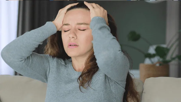A woman in a gray sweater sitting on a couch, holding her head with both hands, eyes closed, appearing overwhelmed or dealing with a headache or anxiety.