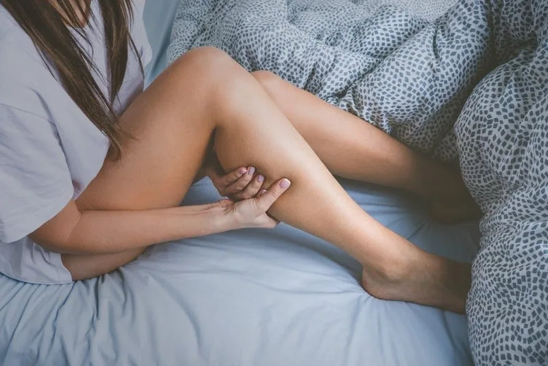 A woman sits on a bed holding her lower leg with both hands, appearing to experience pain or discomfort, possibly due to a muscle cramp or restless leg syndrome.