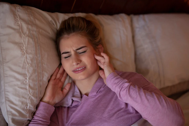 A woman lies in bed wearing a pink hoodie, pressing her hands to her ears with a pained expression, suggesting she is experiencing discomfort or ear pain, possibly due to noise or an ear-related issue.