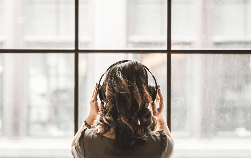 A woman with wavy brown hair wearing headphones, standing near a large window with a cityscape view. She appears to be immersed in music or audio, creating a peaceful and reflective atmosphere.