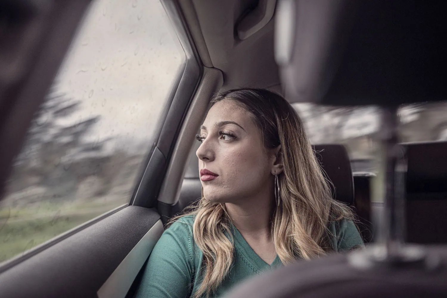 A woman with long blonde hair wearing a green top sits in the backseat of a car, looking thoughtfully out a rain-speckled window on a gloomy day.