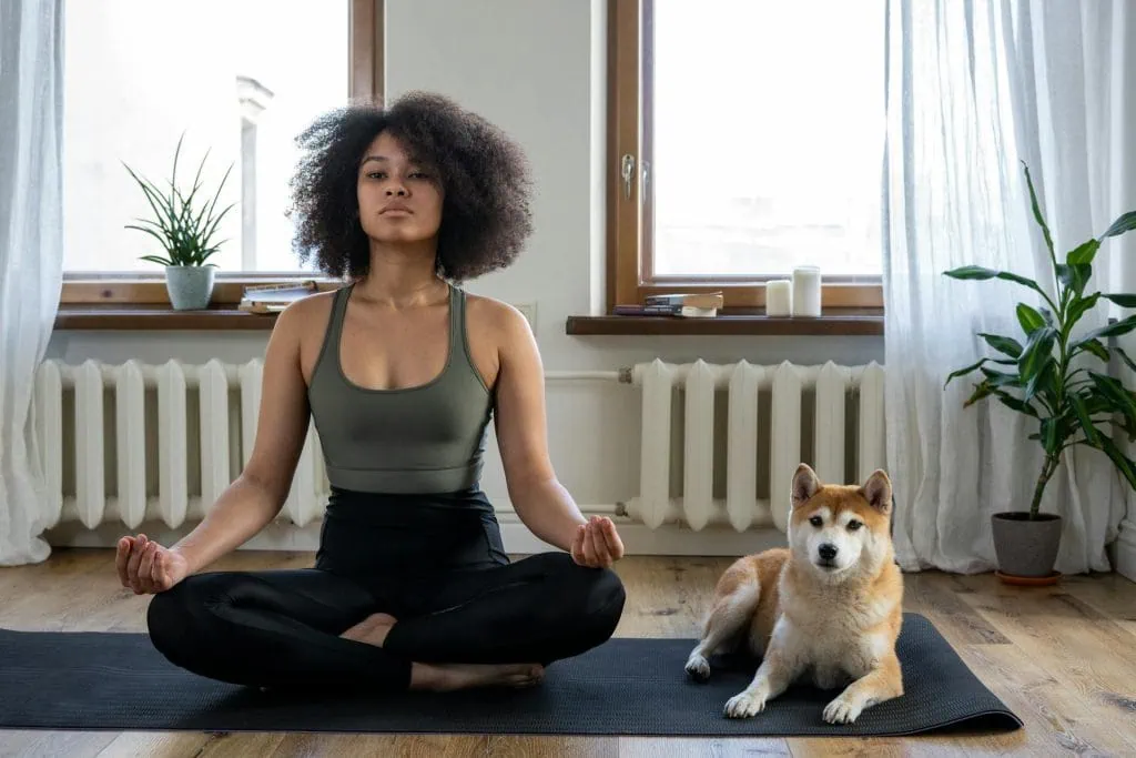 A woman sits cross-legged on the floor in a peaceful meditation pose, accompanied by her relaxed dog beside her, creating a serene moment of mindfulness and companionship.