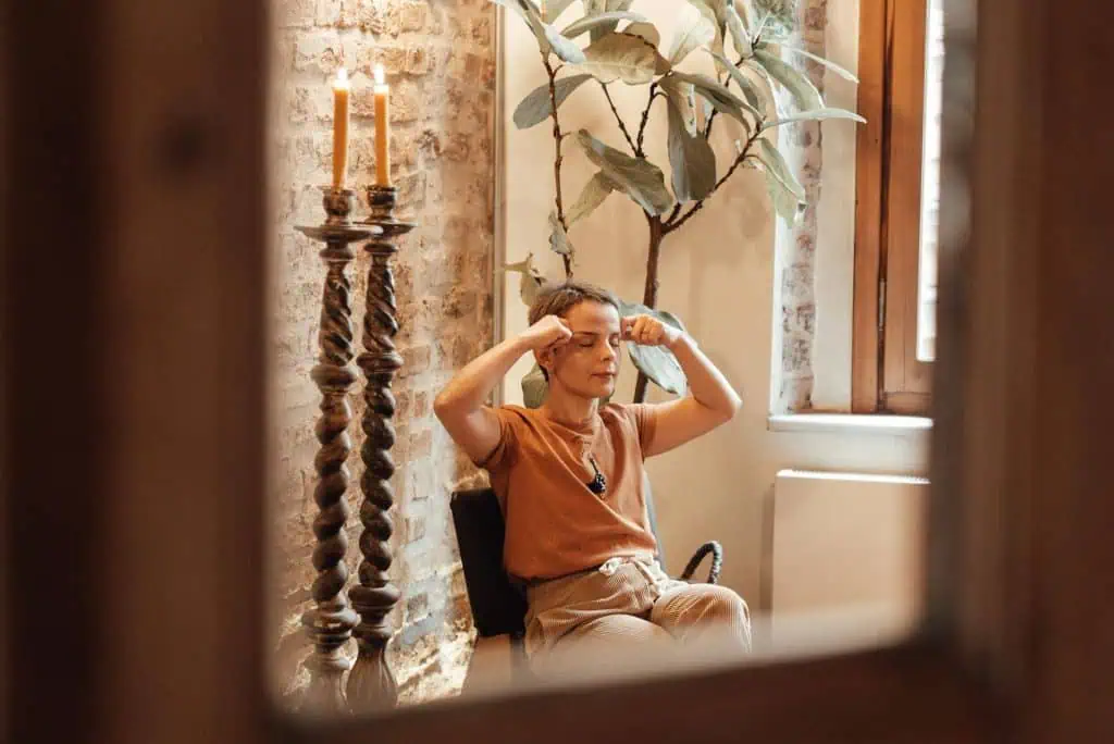 Woman sitting indoors with eyes closed, gently pressing her fingers to her forehead as part of a calming or meditative practice.