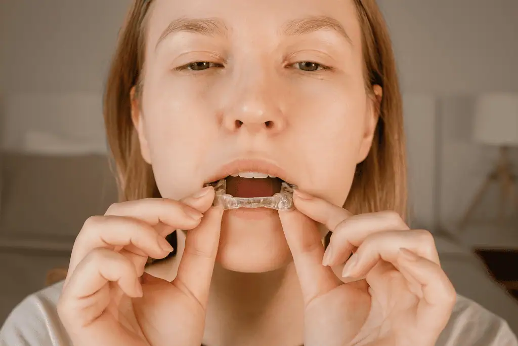 A close-up horizontal image of a woman gently placing a clear dental mouthguard onto her upper teeth, with a softly blurred bedroom background.