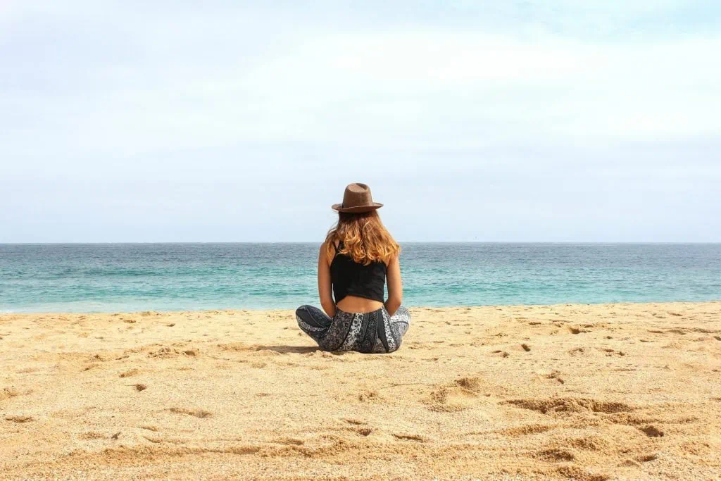 Woman sitting alone on a sandy beach facing the ocean, wearing a hat and relaxed clothing, enjoying a peaceful moment.