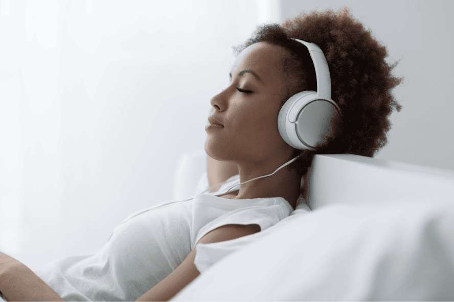 Young woman with curly hair lying on a bed, wearing white headphones and a white shirt, relaxing.