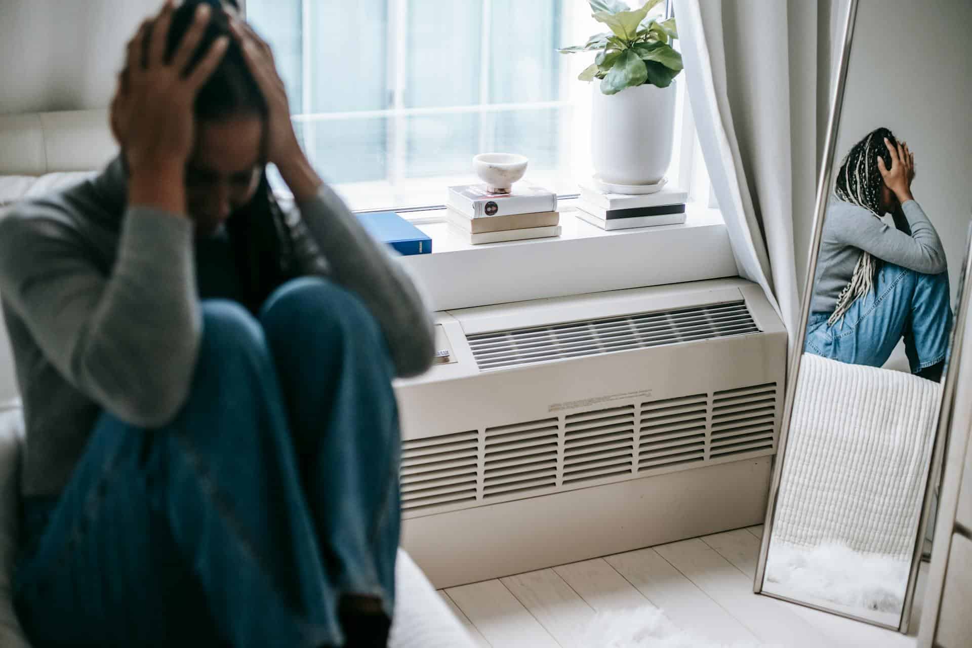 Woman sitting with her head in her hands, reflected in a mirror, showing signs of emotional distress and anxiety.