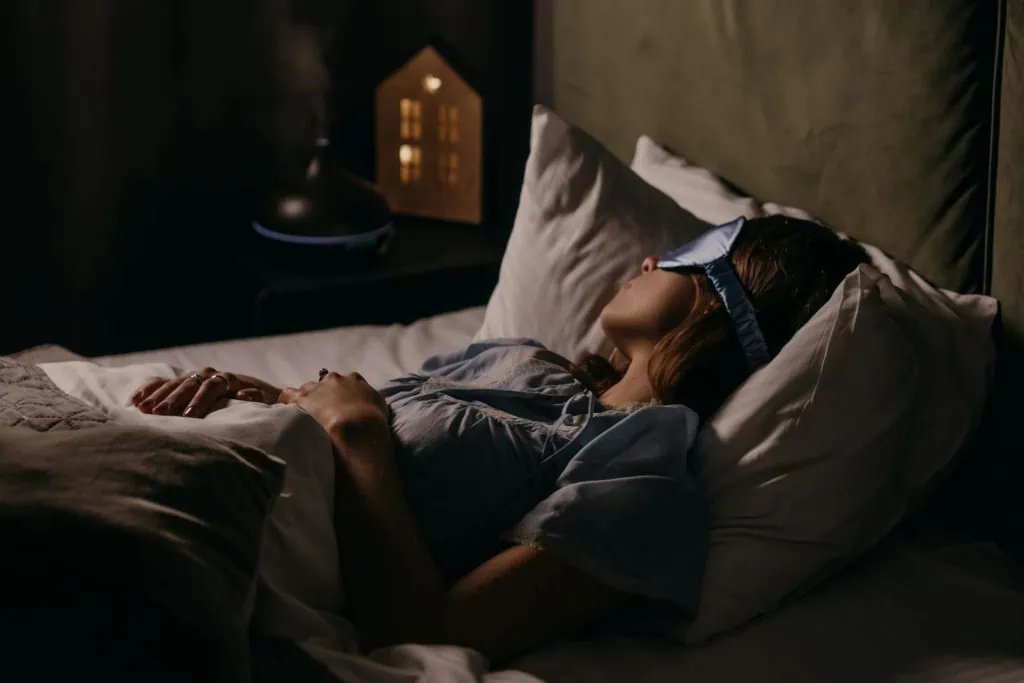 Woman wearing a sleep mask and resting peacefully in bed with an essential oil diffuser in the background