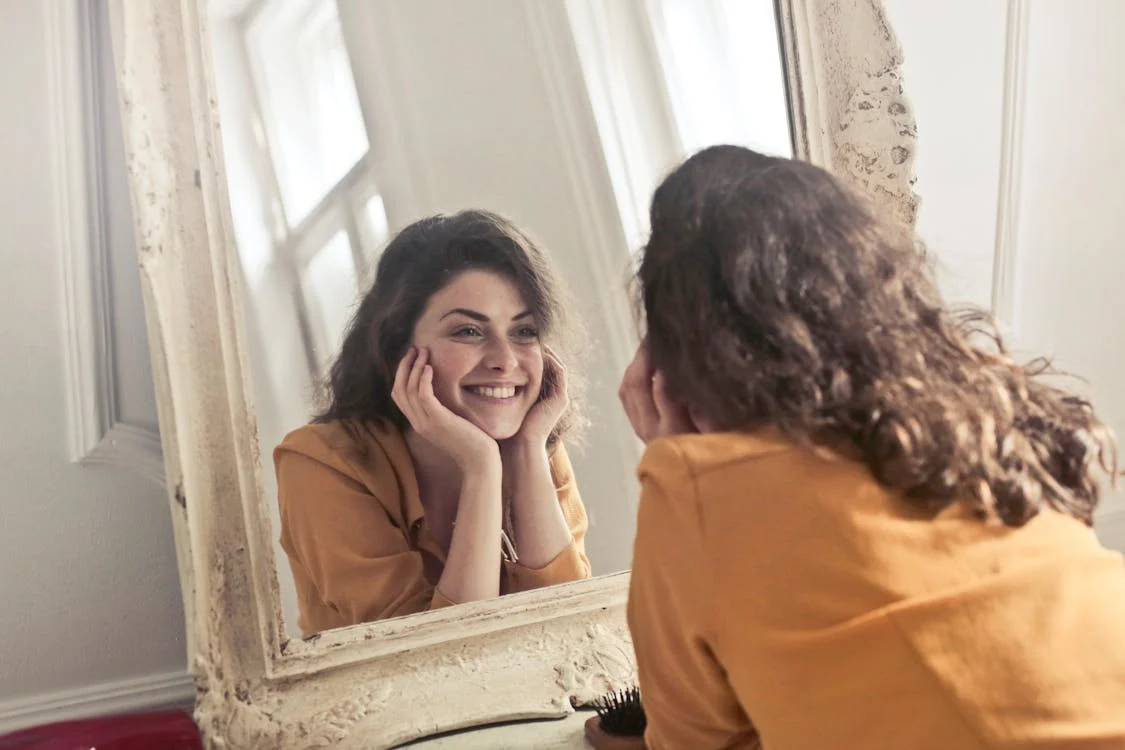A woman with curly hair wearing an orange shirt is smiling and looking at herself in a mirror with her hands resting on her cheeks. The mirror is framed in a vintage, textured white frame, and natural light is coming through a nearby window.