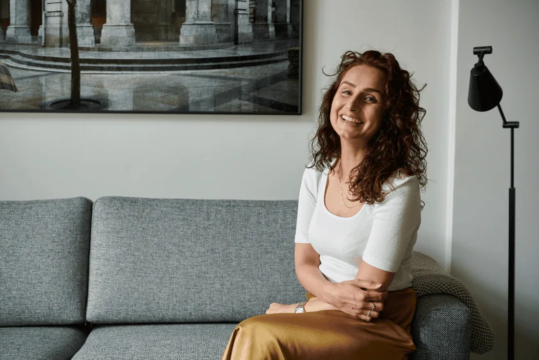 A cheerful woman with curly hair sitting on a gray sofa, smiling and looking relaxed in a modern living room.
