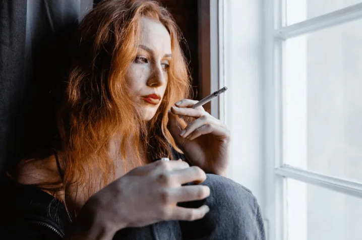 A young woman with red hair and a black jacket smokes a cigarette while sitting by a window, looking contemplative.