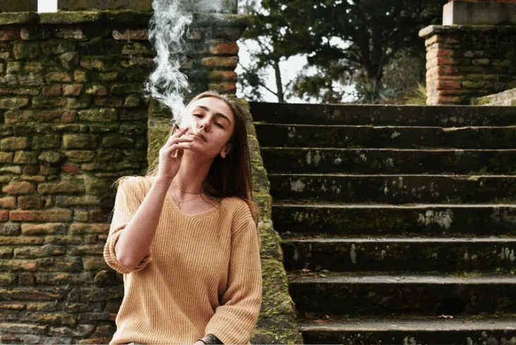 A young woman sitting on outdoor steps, exhaling smoke while smoking a cigarette.