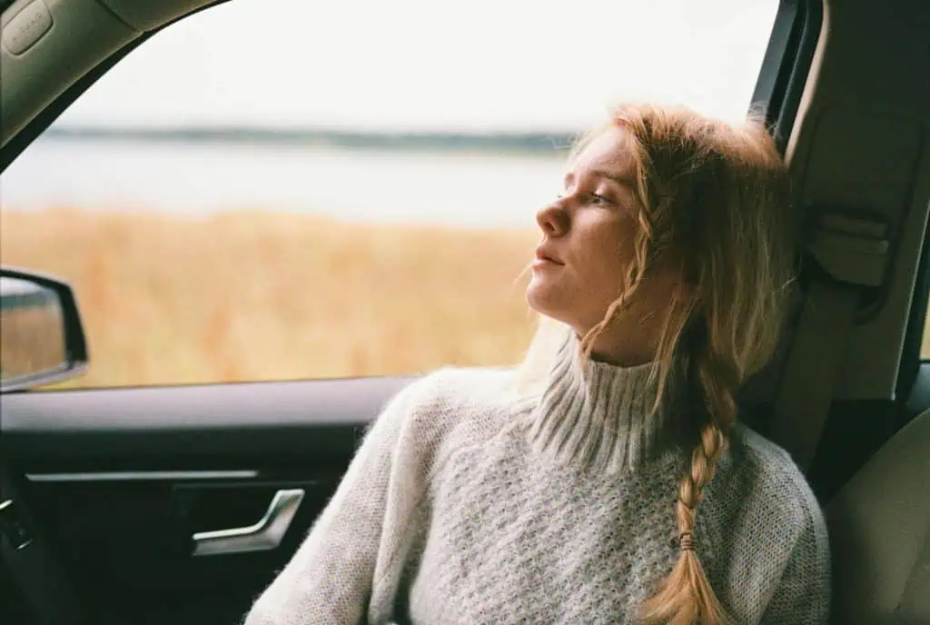 Young woman with a braid in a cozy sweater gazing out of a car window, appearing thoughtful and emotionally distant.
