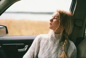Young woman with a braid in a cozy sweater gazing out of a car window, appearing thoughtful and emotionally distant.