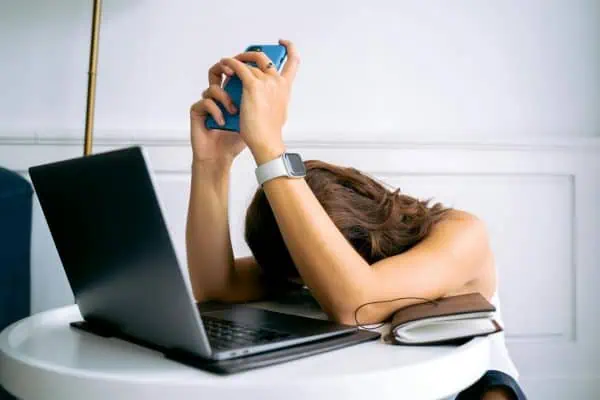 Woman slumped over her desk holding a phone with both hands, showing signs of stress and digital burnout next to a laptop and notebook.