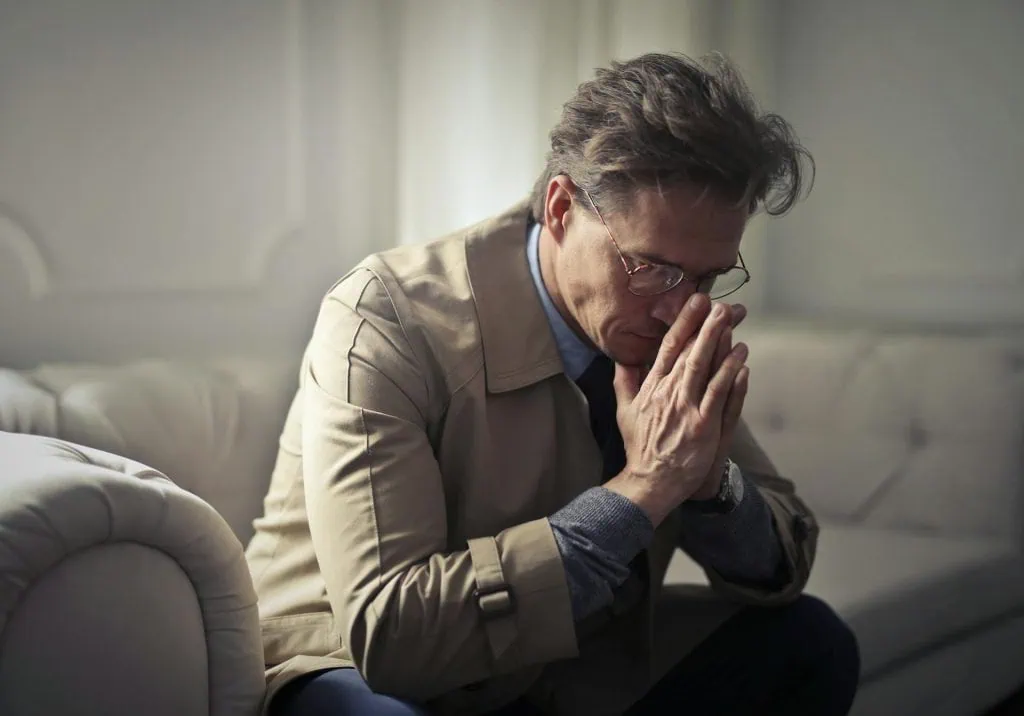 Worried man in a beige coat sitting on a couch with hands clasped near his face, appearing deep in thought or prayer.