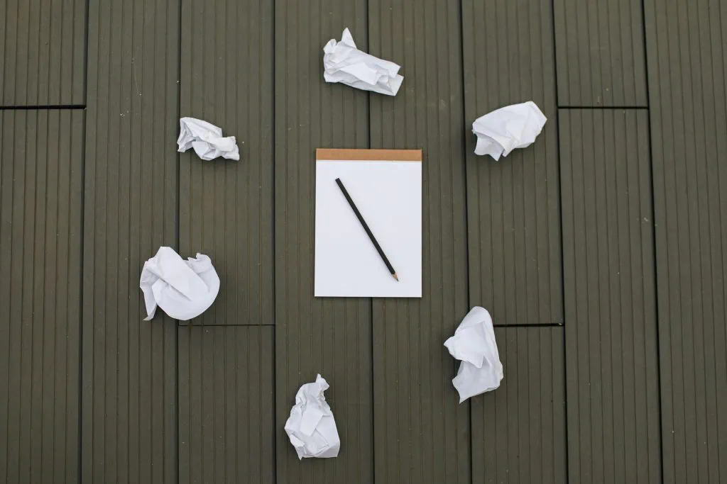 Blank notepad with a pencil surrounded by crumpled paper balls on a wooden surface, symbolizing writer’s block or creative frustration.