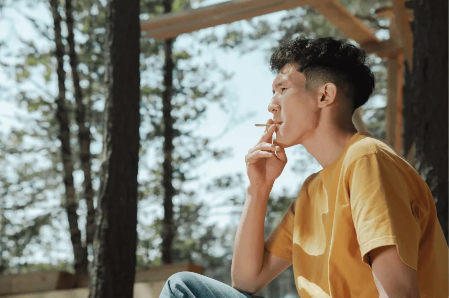 Young man wearing a yellow t-shirt smoking a cigarette while sitting outdoors near trees.