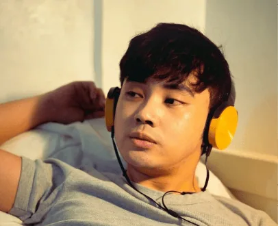 Young man reclining on a bed while wearing yellow headphones, gazing thoughtfully into the distance.