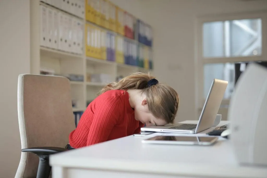 Young woman exhausted at work, resting her head on the desk in front of a laptop in an office environment.